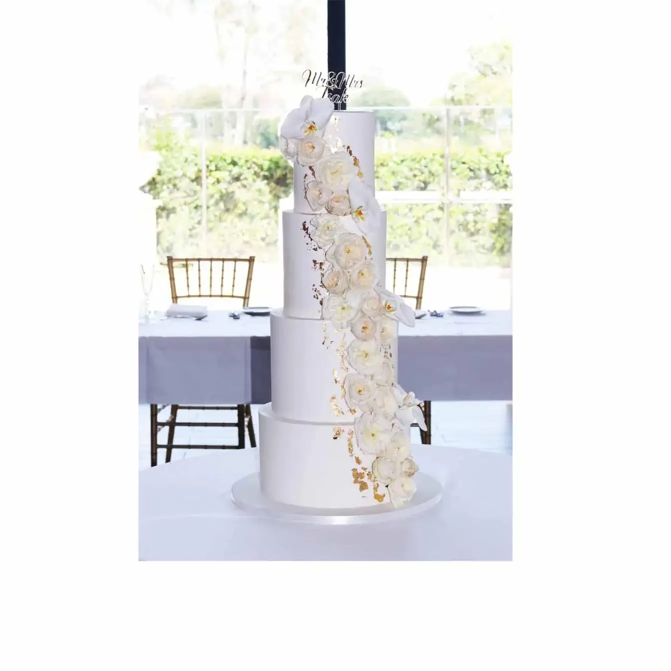 Elegant Gold and Floral Wedding Cake - A four-tier white cake with cascading gold leafing, David Austin roses, white orchids, and a custom name cake topper, a stunning centerpiece for a romantic and elegant wedding celebration.